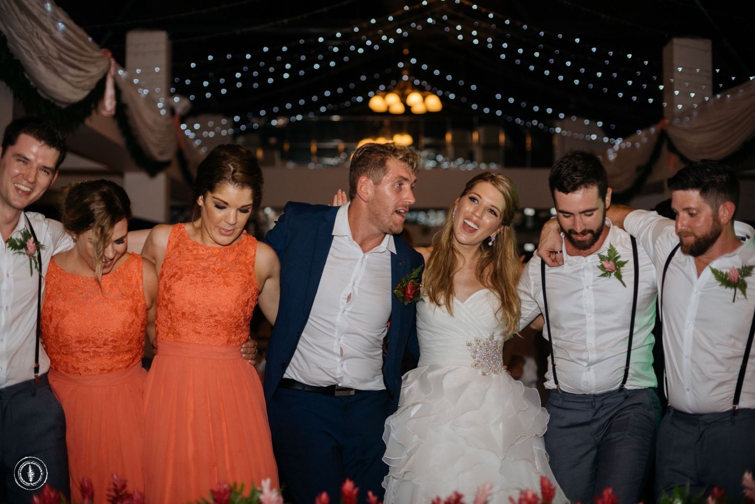 Wedding party in coral dresses and suspenders posing together under twinkling lights.
