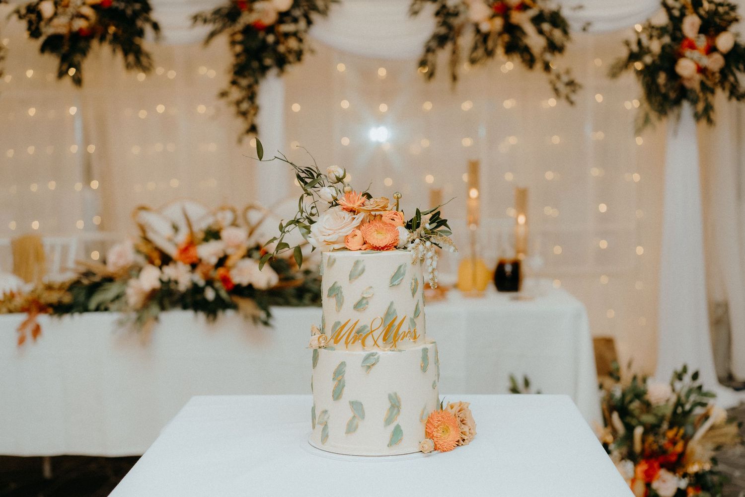 Elegant three-tier white wedding cake decorated with peach flowers and greenery against a lit backdrop.