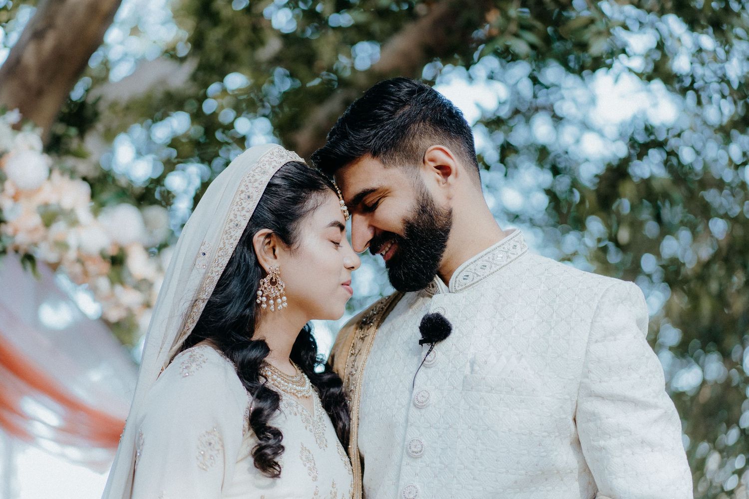 South Asian bride and groom in white traditional attire share an intimate forehead touch outdoors.