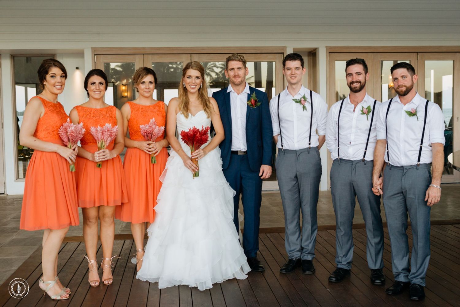Wedding party poses in coral and gray attire on wooden deck.