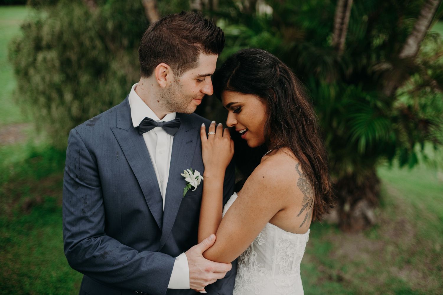 Intimate moment between couple in formal wedding attire surrounded by tropical foliage.