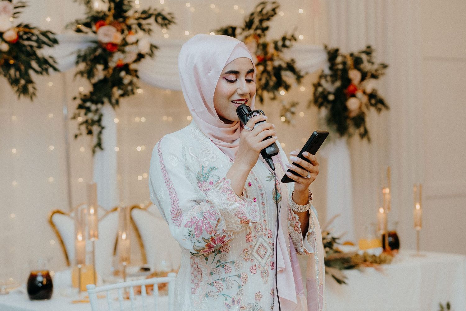 Person in white embroidered outfit speaks into microphone during wedding celebration.