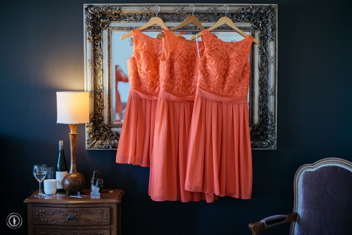 Three coral colored bridesmaid dresses hanging on display against dark wall with ornate frame.