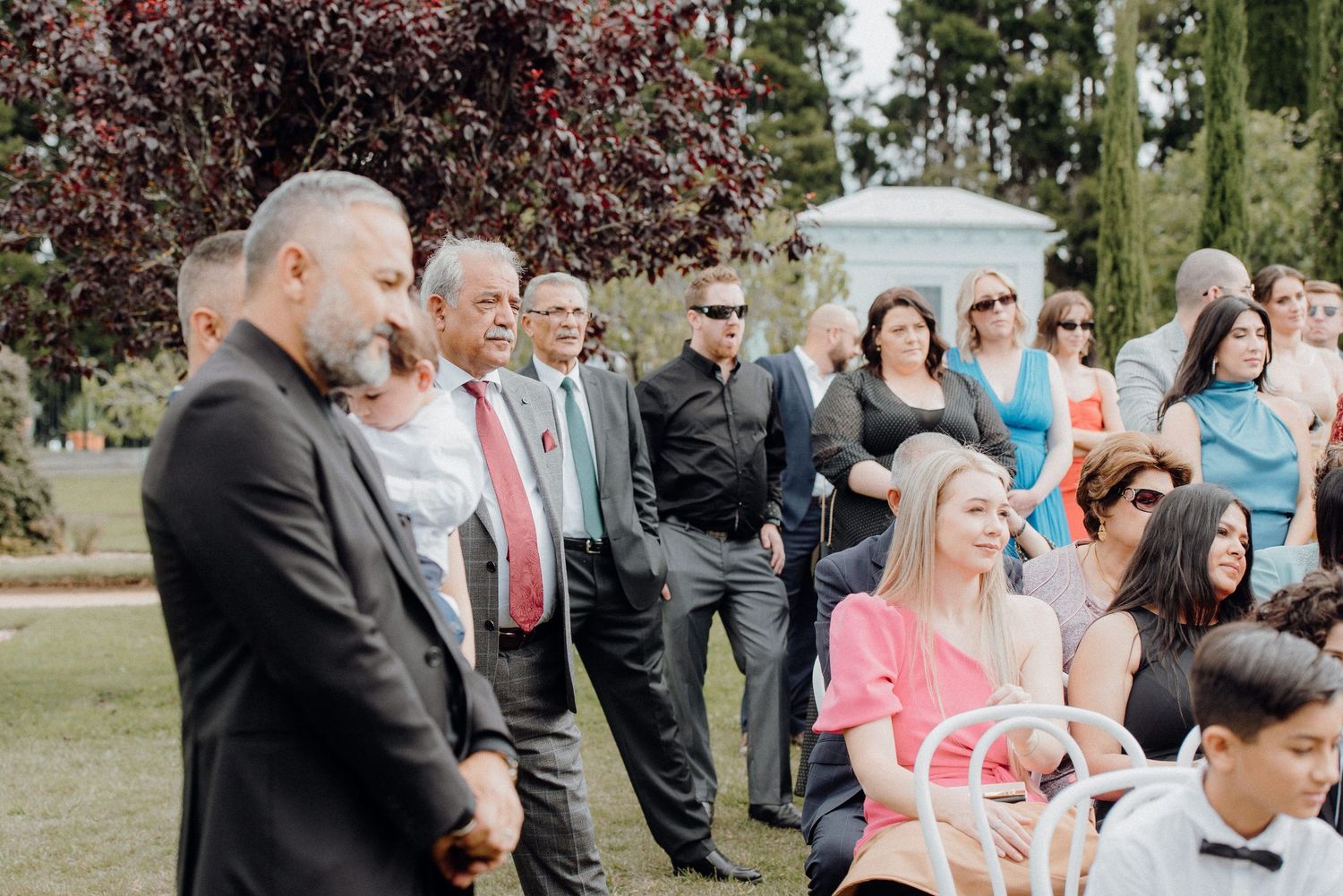 Wedding guests and attendees gather outdoors during ceremony surrounded by natural landscaping.