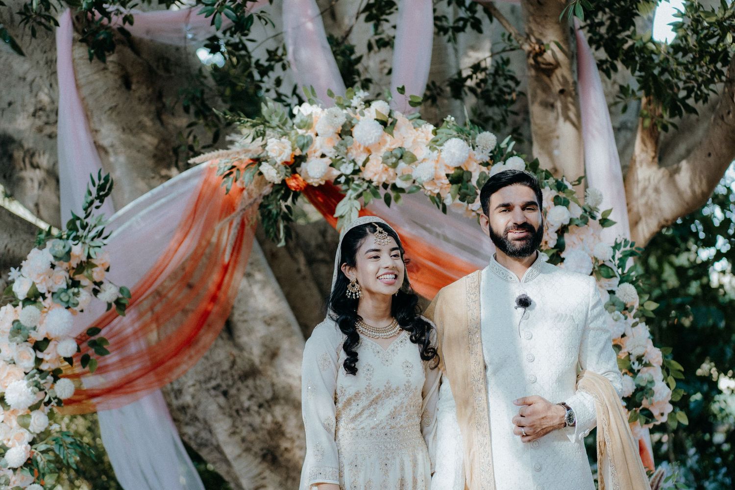 Elegant couple in white traditional attire stand together under floral wedding arch with coral accents.
