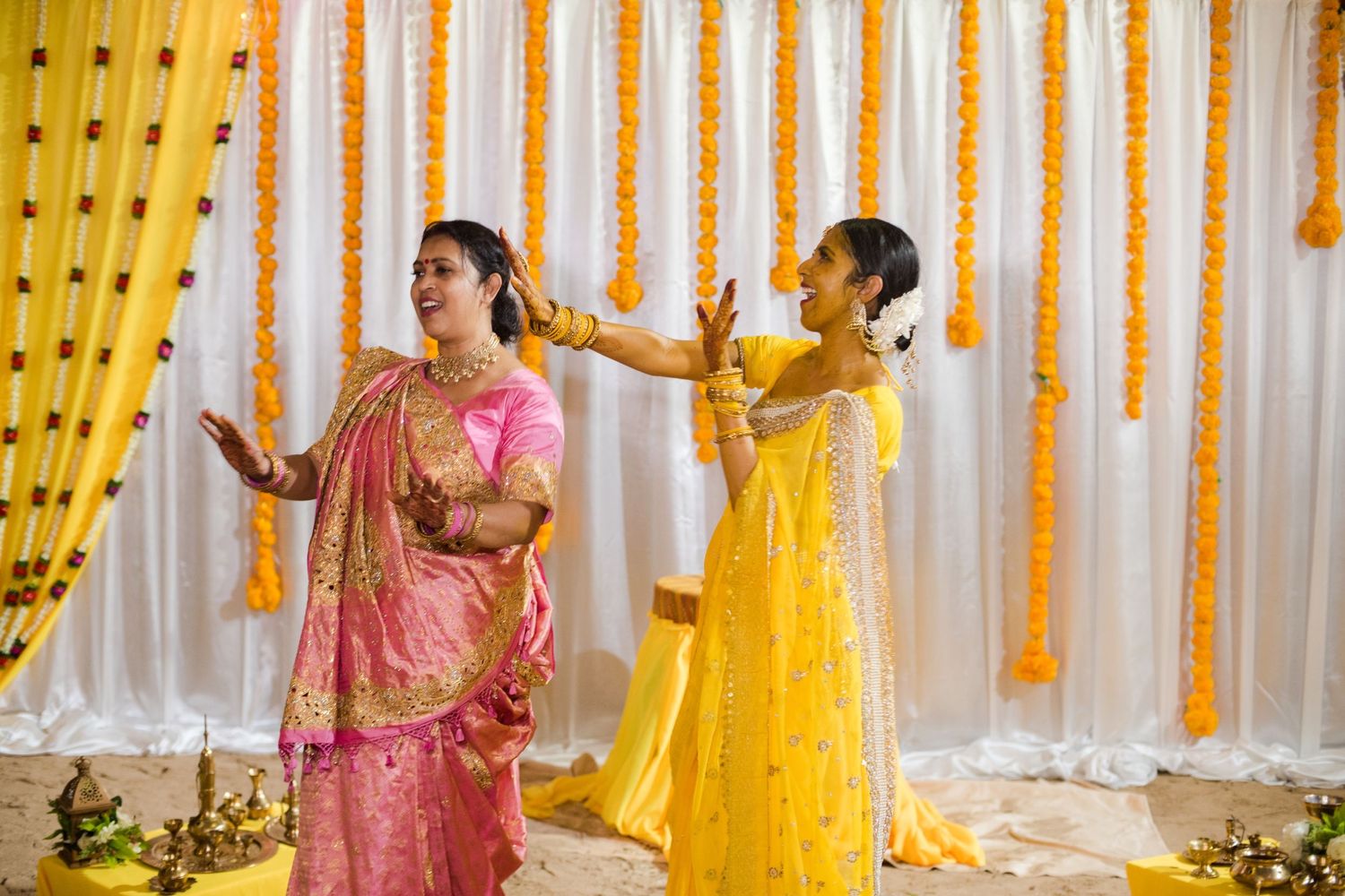 Two women in colorful traditional attire performing dance moves against marigold decorated backdrop.