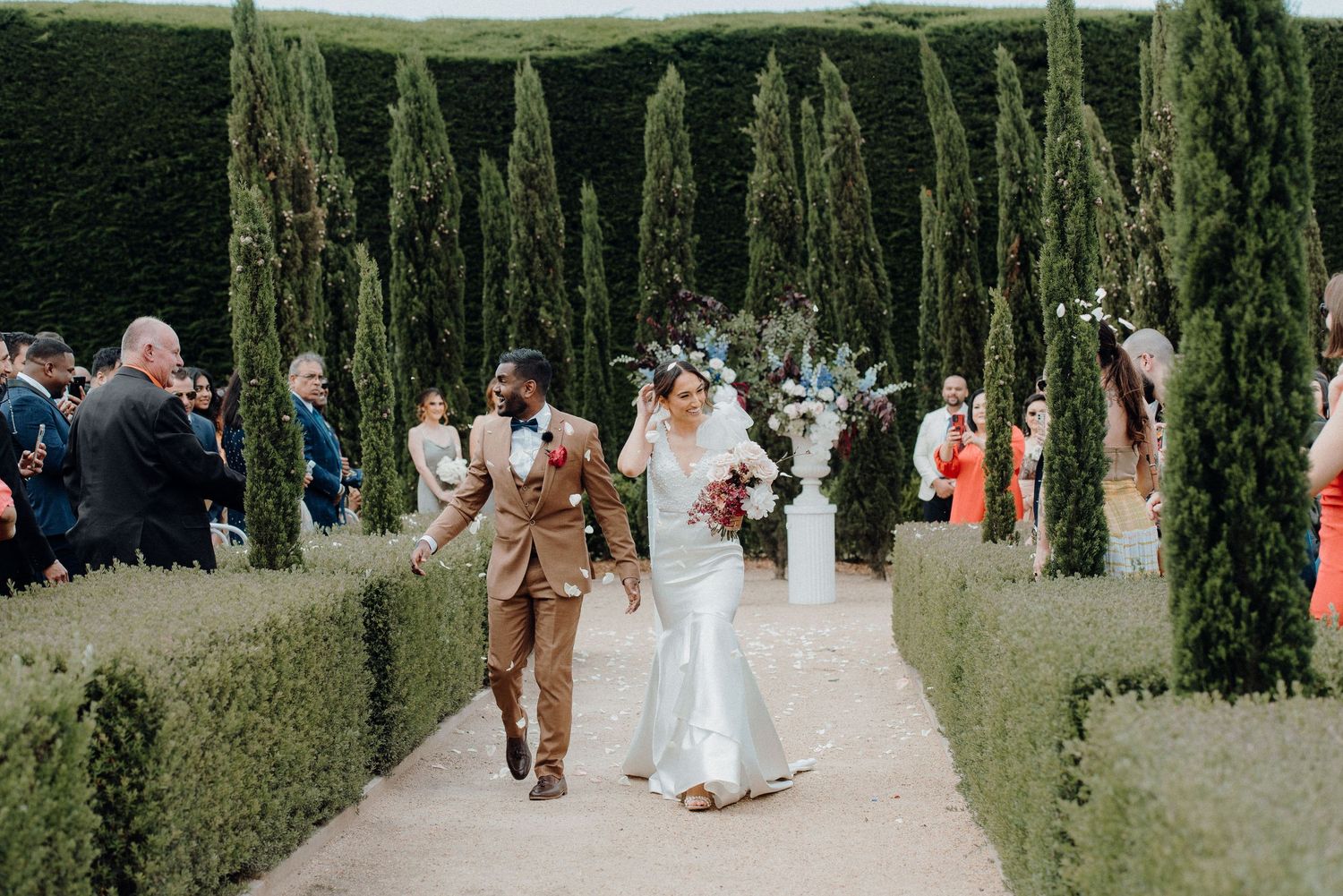 A couple walks down a garden path lined with tall cypress trees and manicured hedges during a wedding ceremony.
