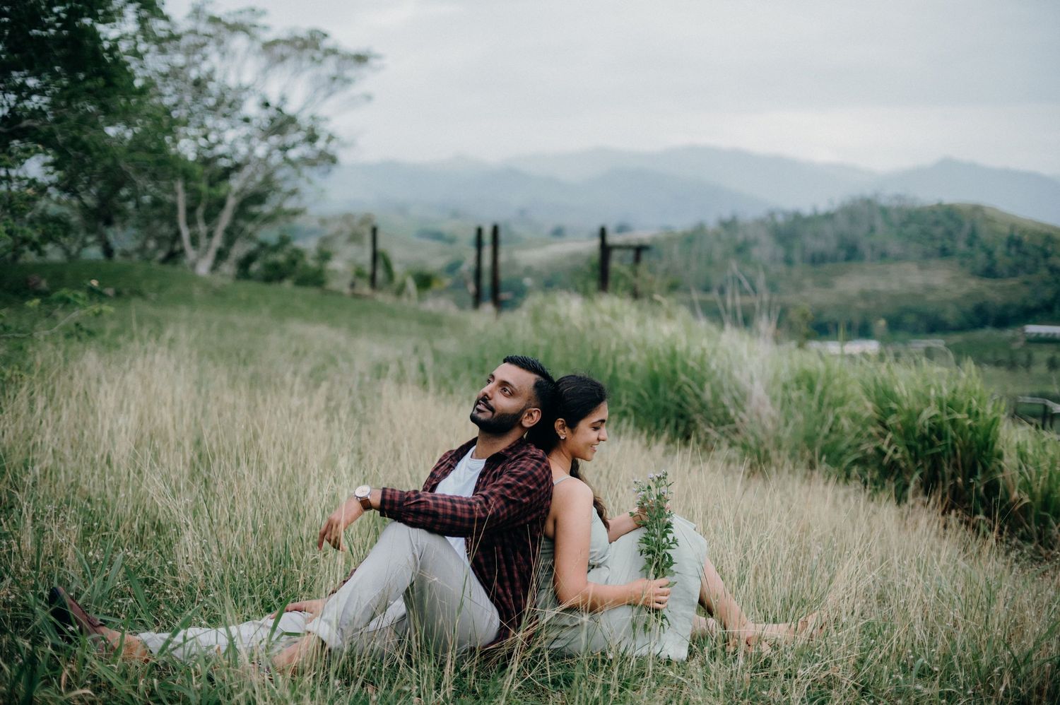 A couple sits back-to-back in tall grass with mountains in the distance.