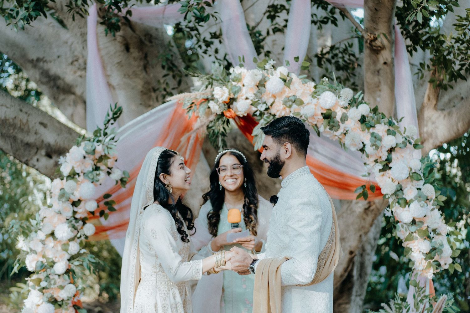 Wedding officiant conducts ceremony for couple under floral arch with white and coral decorations.