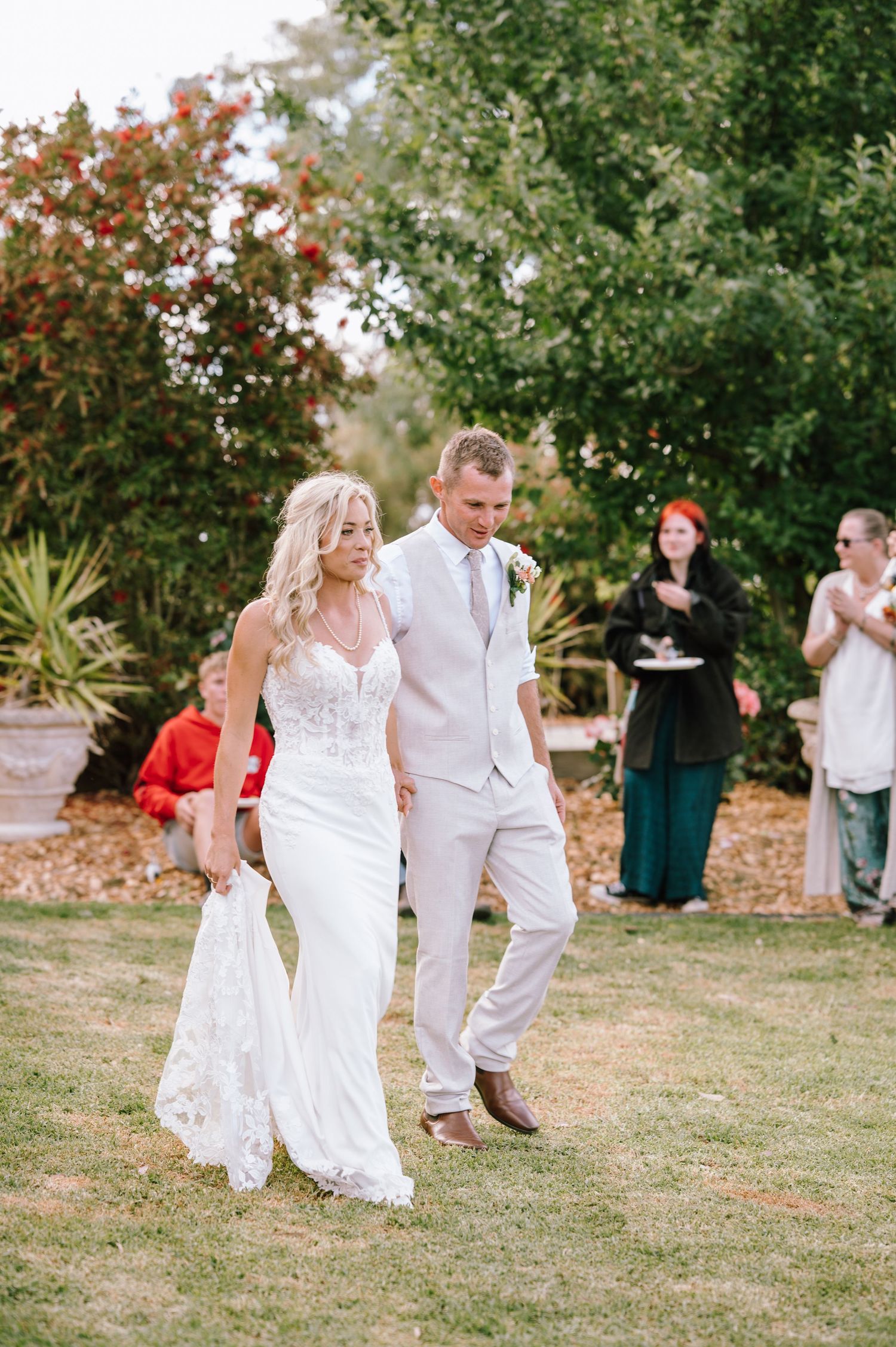 Blonde bride in fitted lace gown walks with groom in light suit at outdoor wedding.
