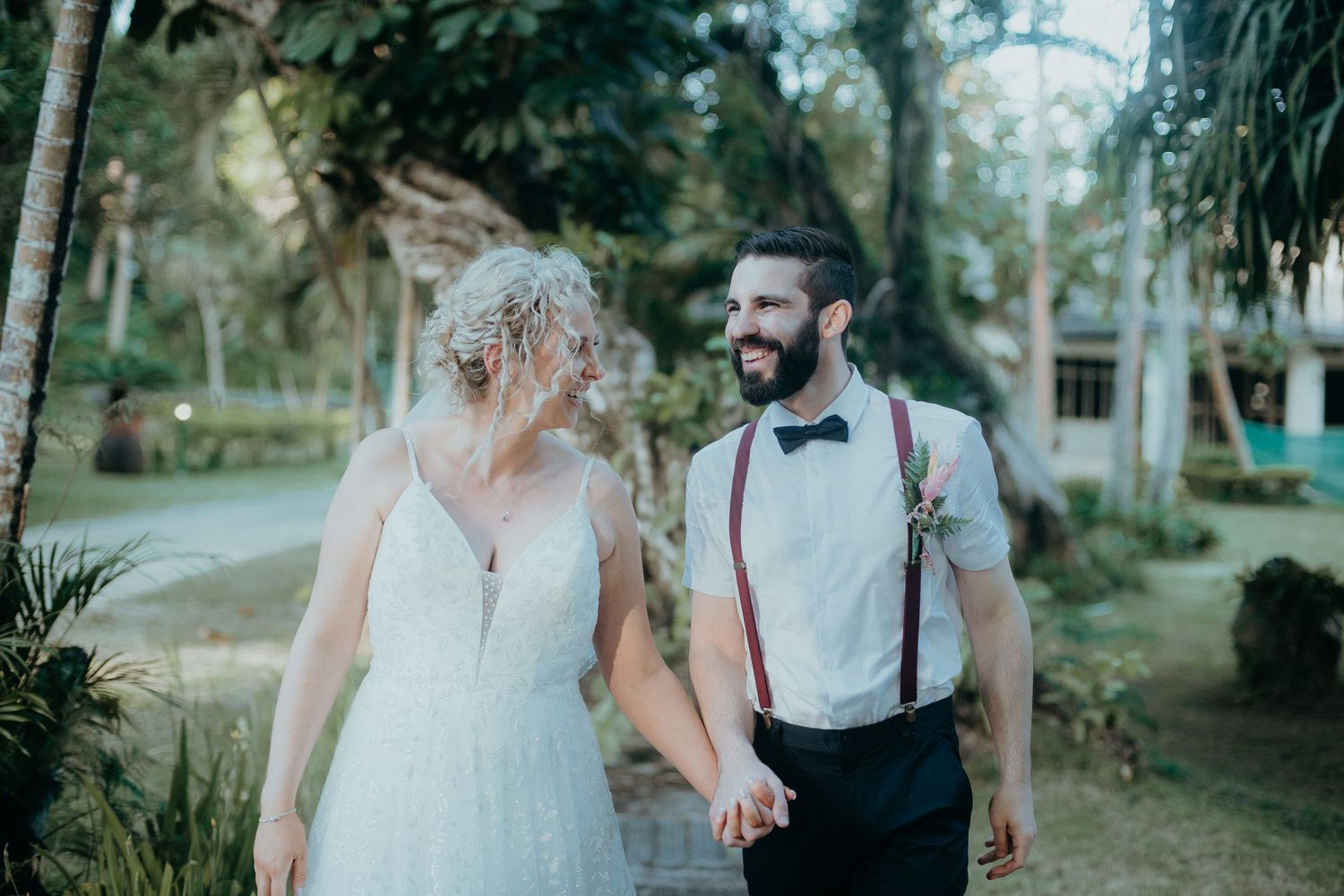 Bride in lace gown and groom in suspenders hold hands walking through a lush tropical garden setting.