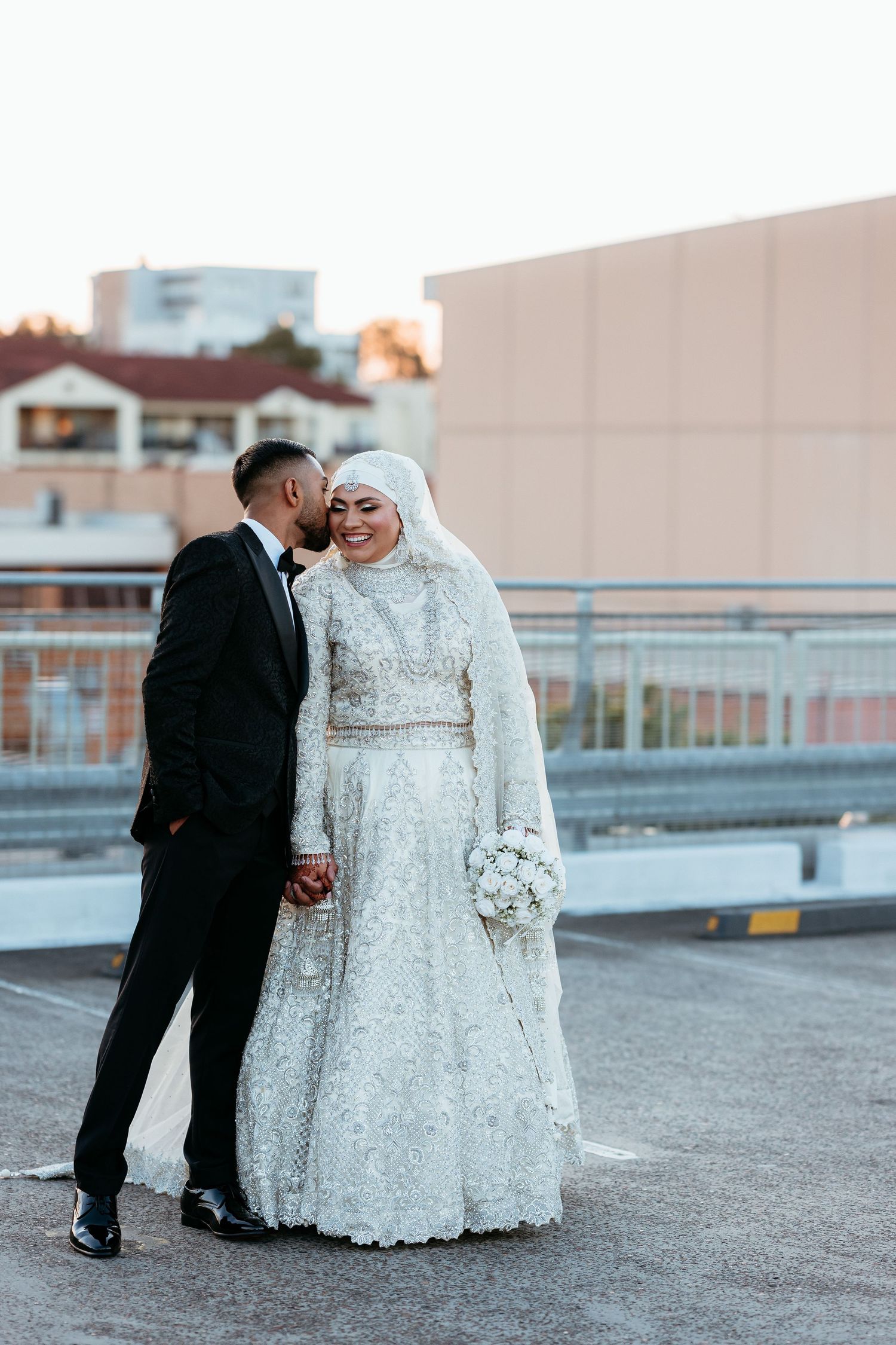 Groom kissing bride on cheek, bride in white lace hijab gown holding bouquet on rooftop.