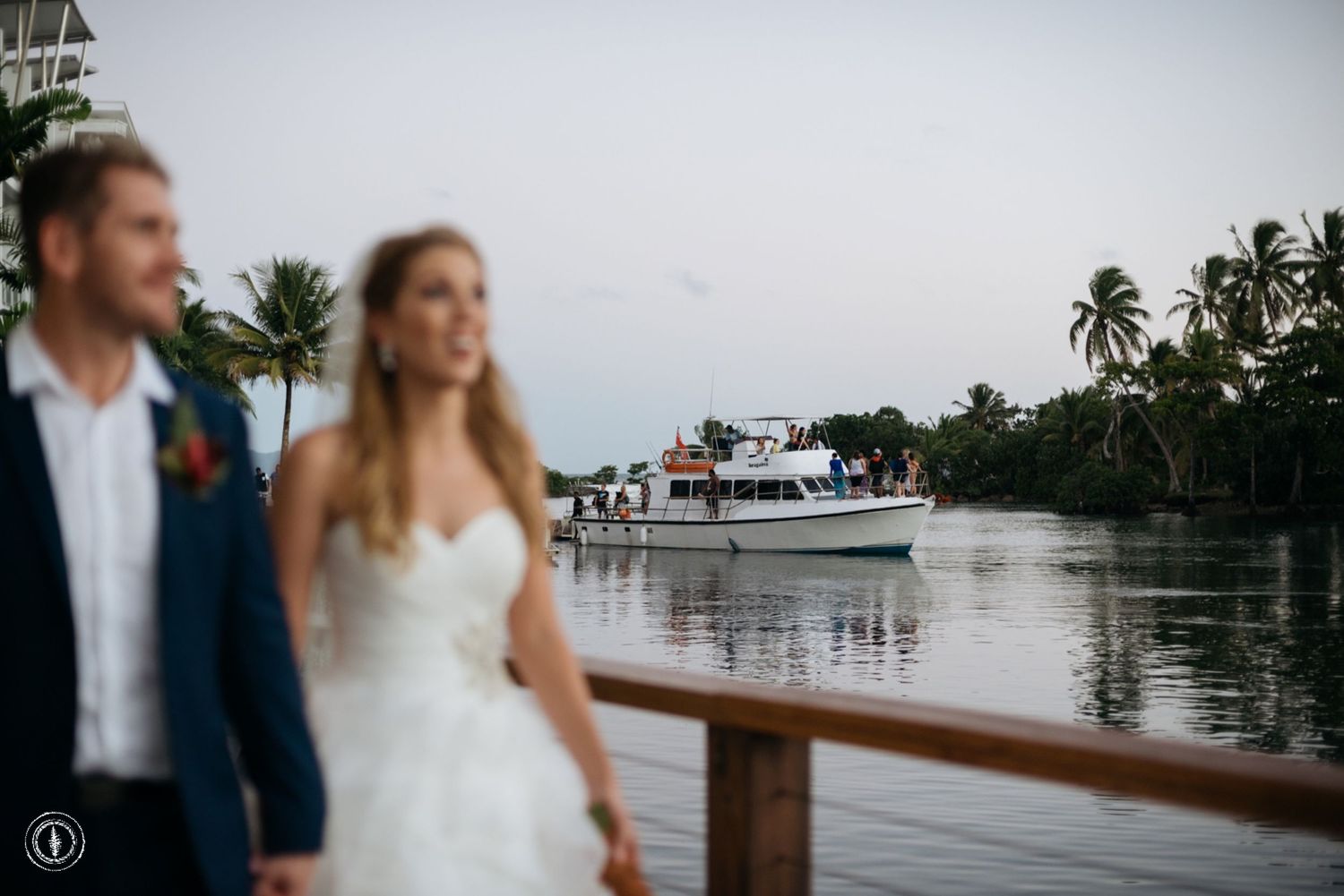 Candid wedding photo by the water with boat and palm trees in background.