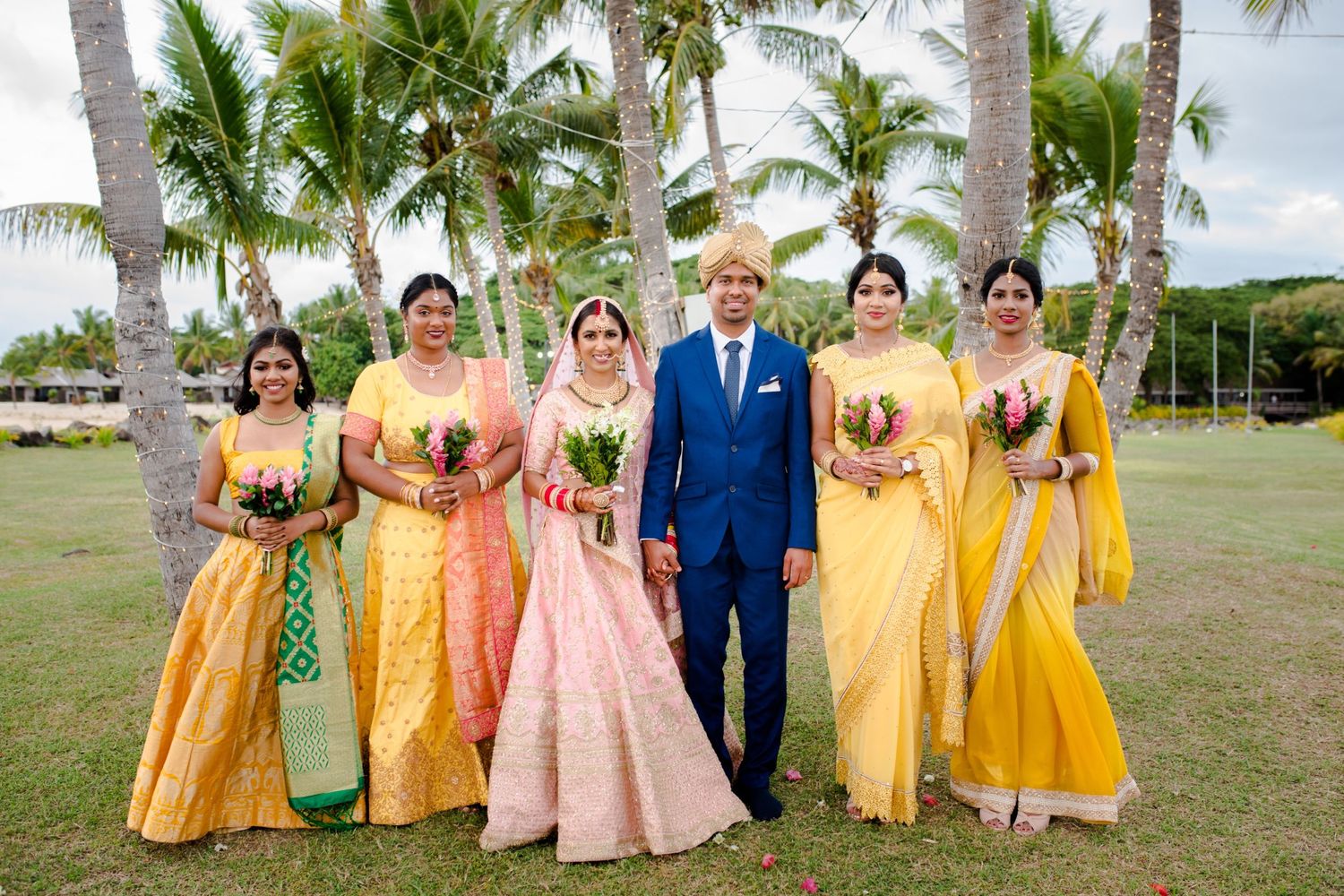 Wedding party in coordinated yellow and pink attire poses together under tropical palm trees.