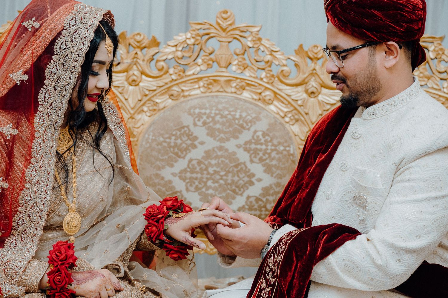A couple in traditional South Asian wedding attire exchanges rings on an ornate golden throne.