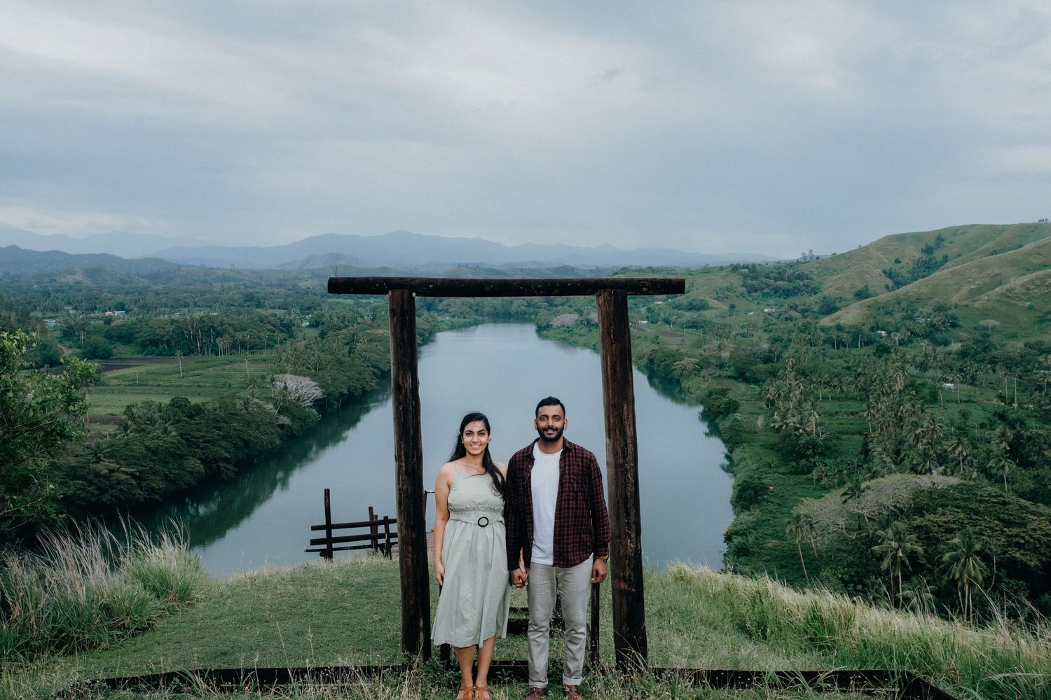 A couple stands beneath a wooden arch overlooking a scenic river valley and rolling hills.
