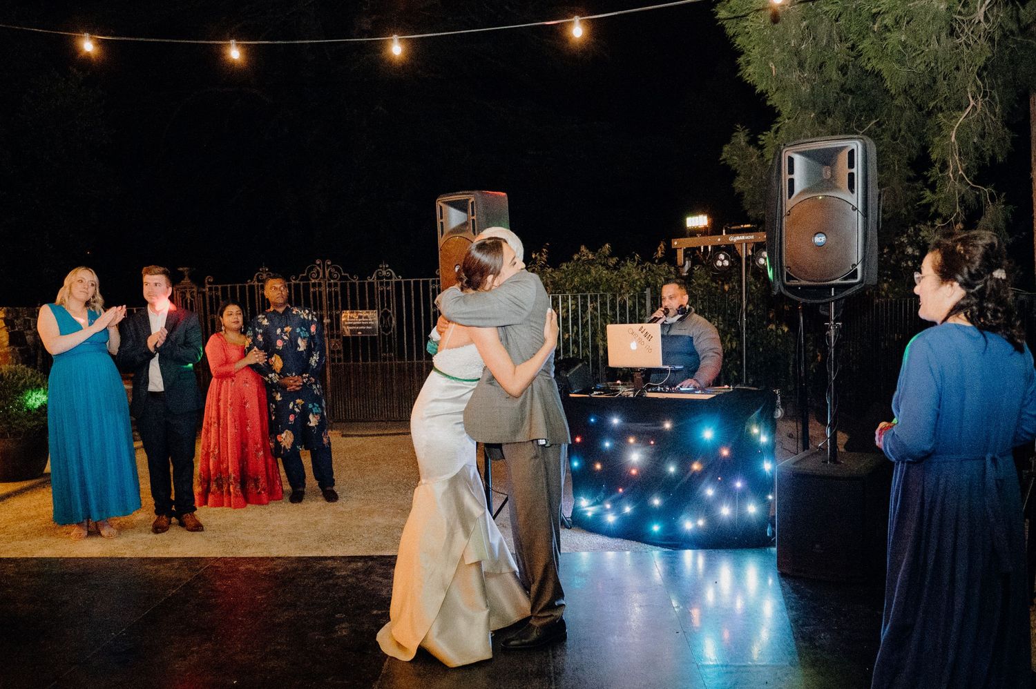 Couple slow dancing at night under string lights with a DJ booth in the background.