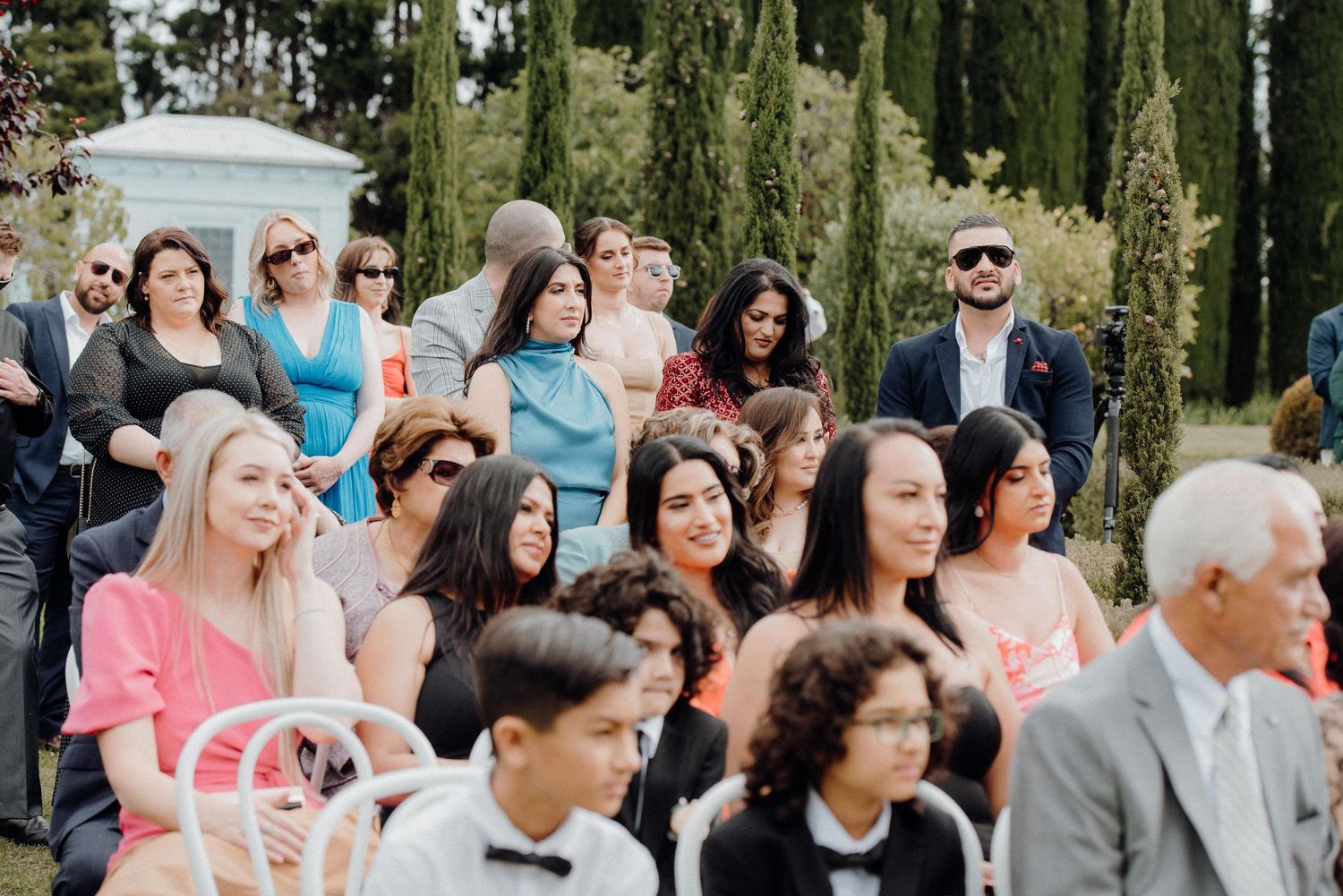 Wedding guests seated in white chairs during outdoor ceremony surrounded by cypress trees.