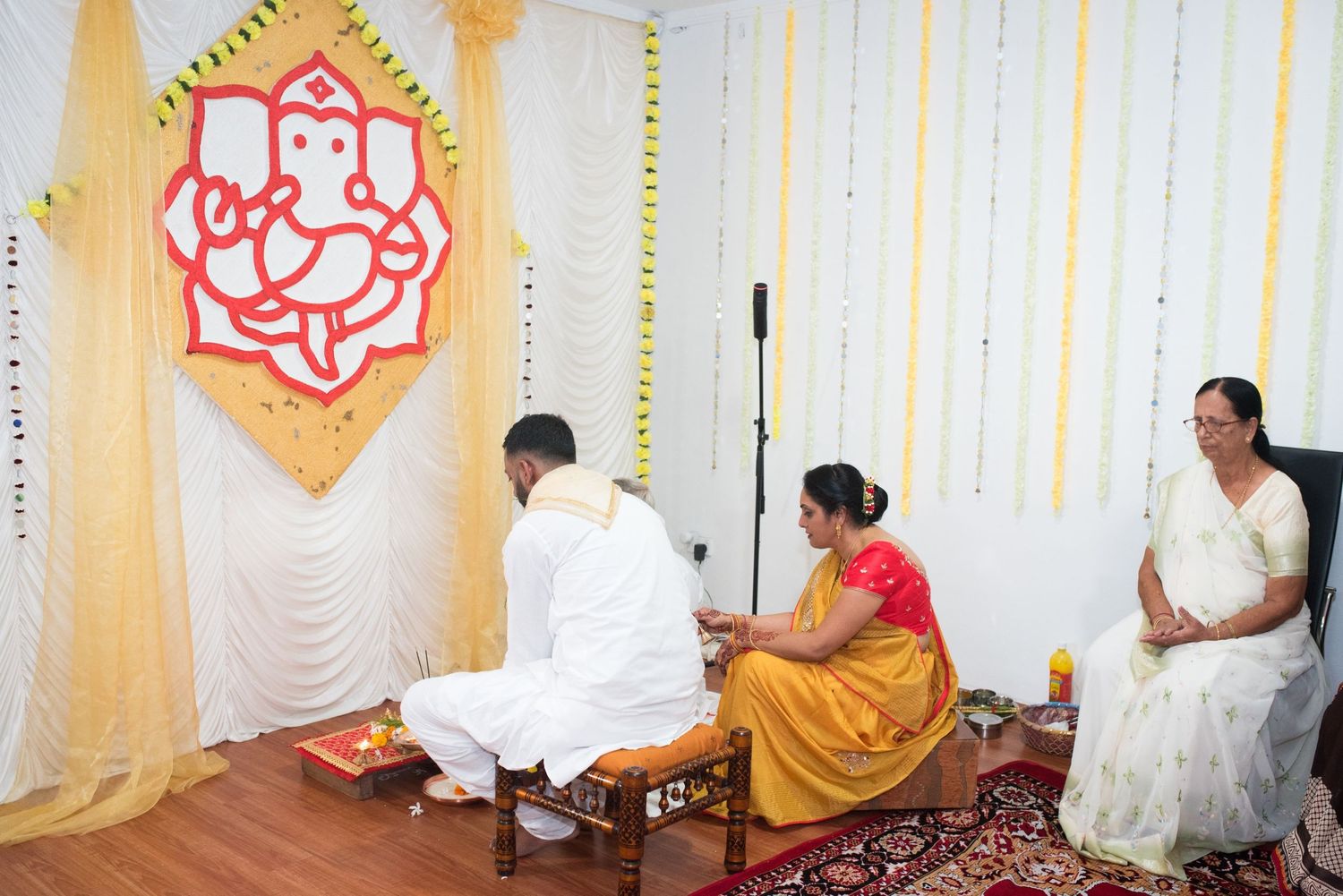 An indoor ceremony setup with a Ganesh wall decoration and traditional Indian decor featuring yellow and white drapes.