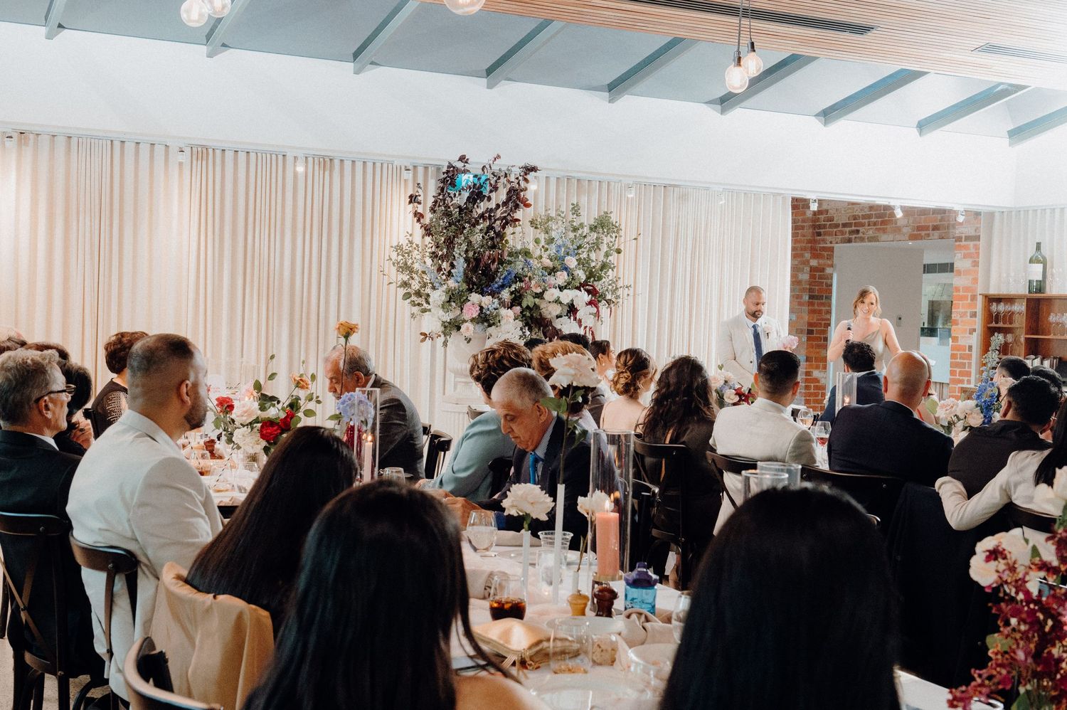 Elegant wedding reception with a large floral centerpiece in a bright venue filled with dining guests.