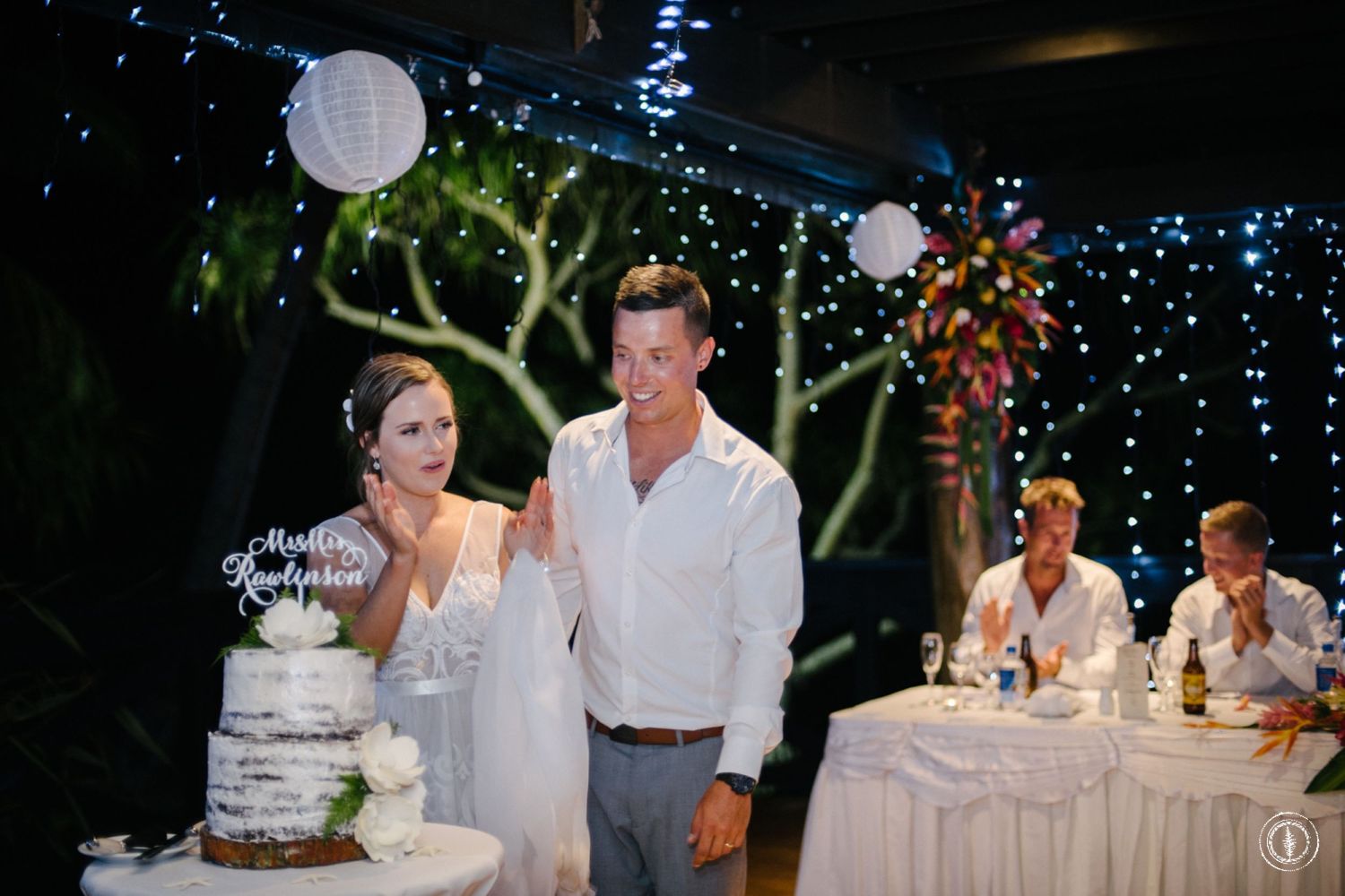 Couple performing cake cutting ceremony at evening reception under string lights.