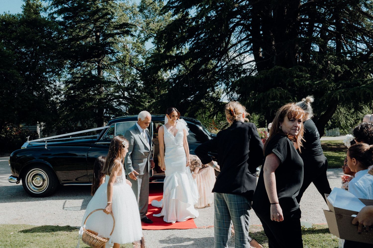 Wedding party gathers around a classic black vintage car on red carpet.