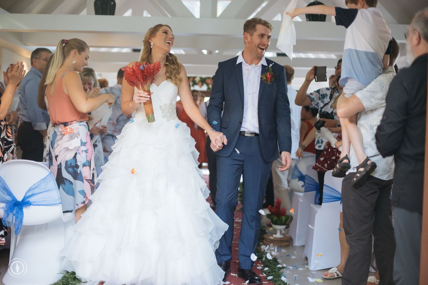 A wedding celebration in a bright indoor venue with floral decorations and white chair covers.