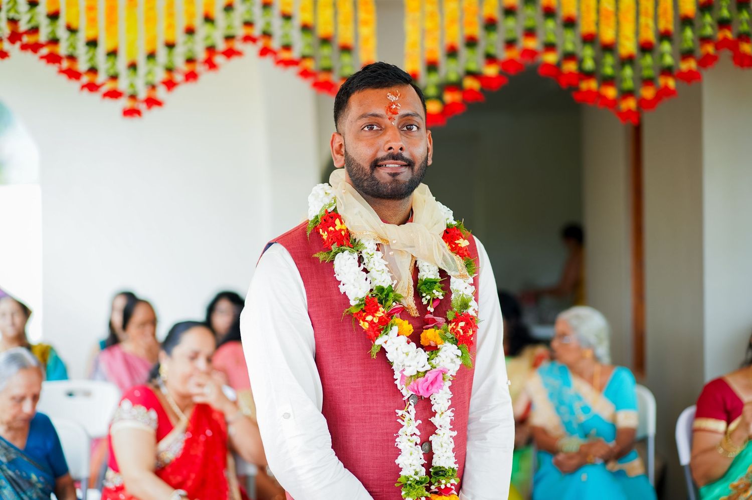 Wedding participant in red vest and white floral garlands during traditional ceremony.
