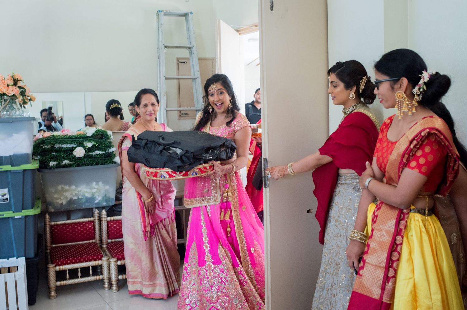 Women in vibrant traditional attire carry garments during wedding preparations.