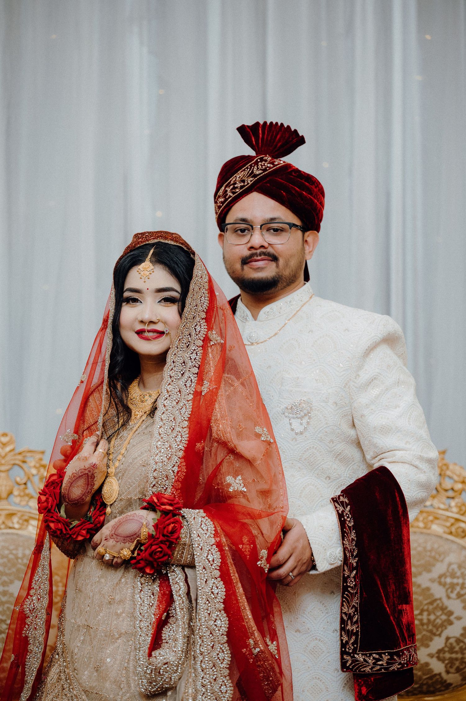 Bride in red and gold wedding attire poses with groom in white sherwani against light backdrop.
