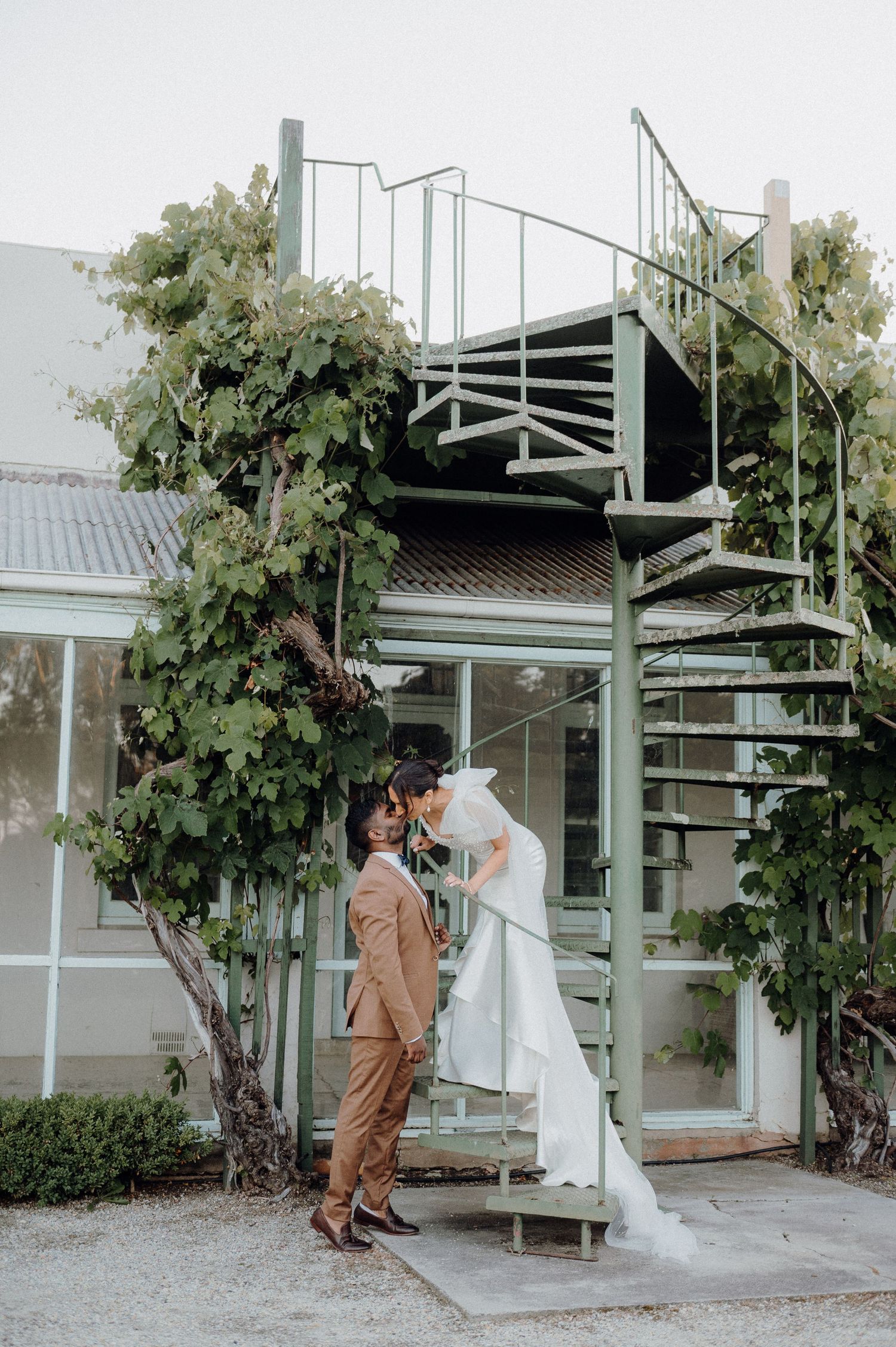 Couple poses on spiral staircase covered in climbing vines during wedding photoshoot.