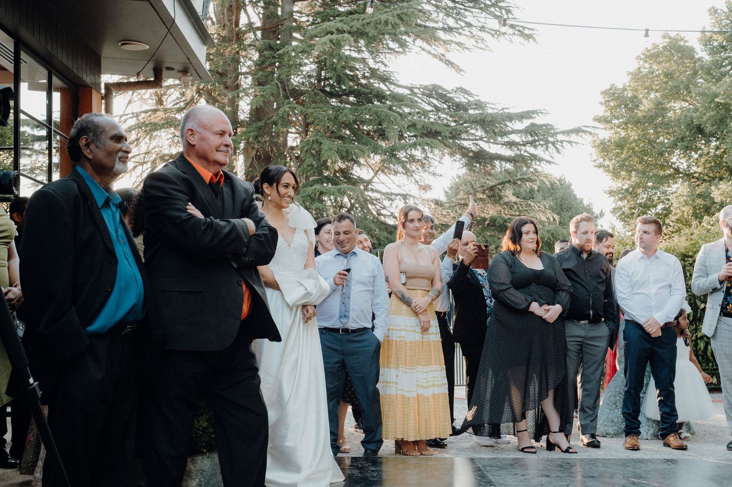 Wedding guests gathered outside under tall trees watching a celebration.