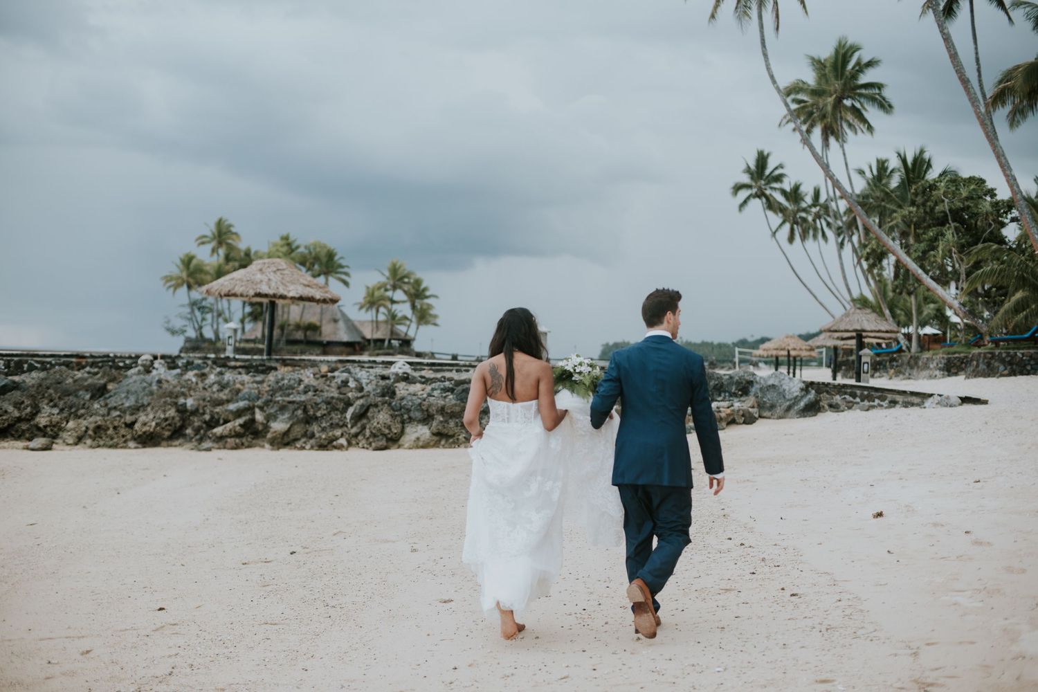 Couple walking hand in hand on a tropical beach with palm trees and rocky coastline.