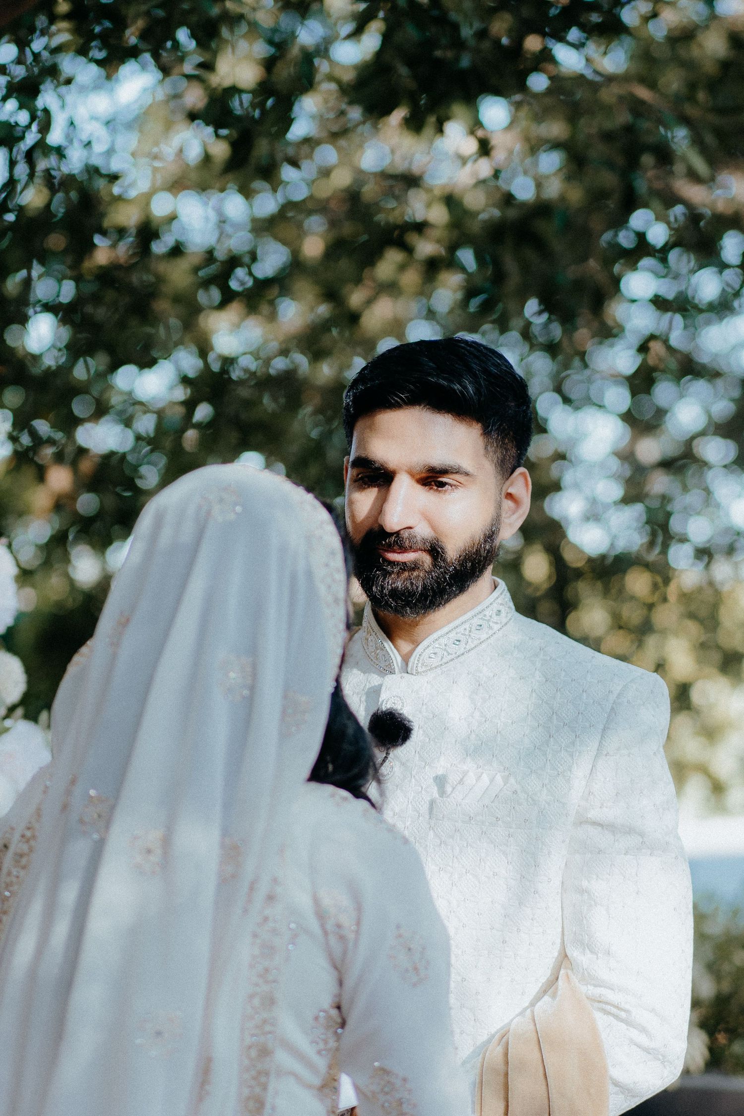 A groom in a white traditional outfit stands during an outdoor wedding ceremony.