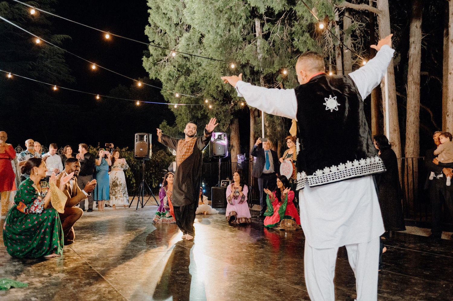 Traditional folk dancers performing outdoors under string lights with tall trees in the background.