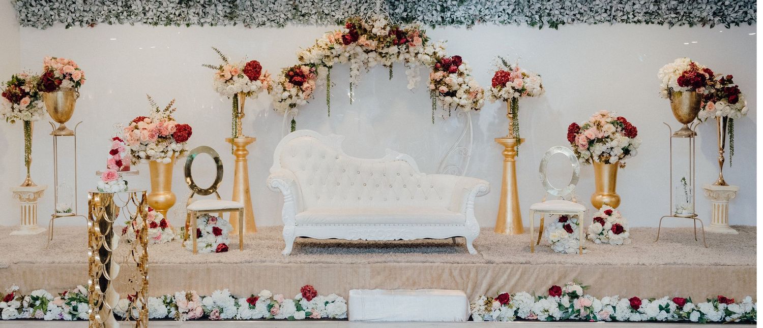 Elegant wedding stage setup with white sofa, gold pillars, and pink and red floral arrangements.