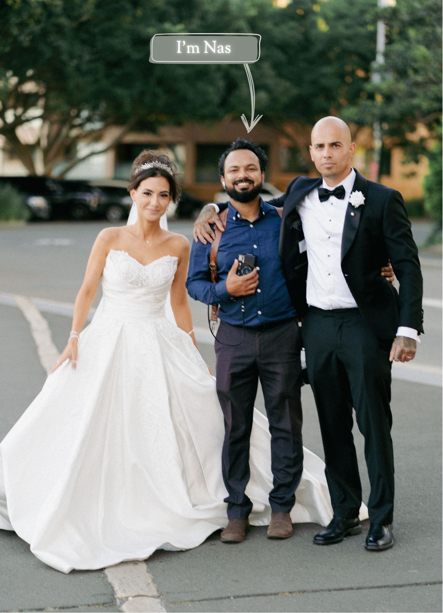 Wedding photographer Nas poses with a bride in a white gown and groom in a tuxedo outdoors.