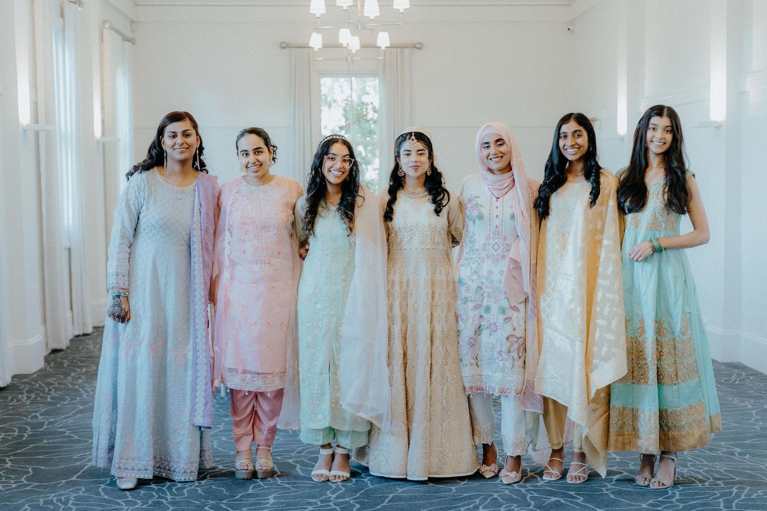A group portrait of wedding guests in pastel traditional South Asian attire against a white backdrop.