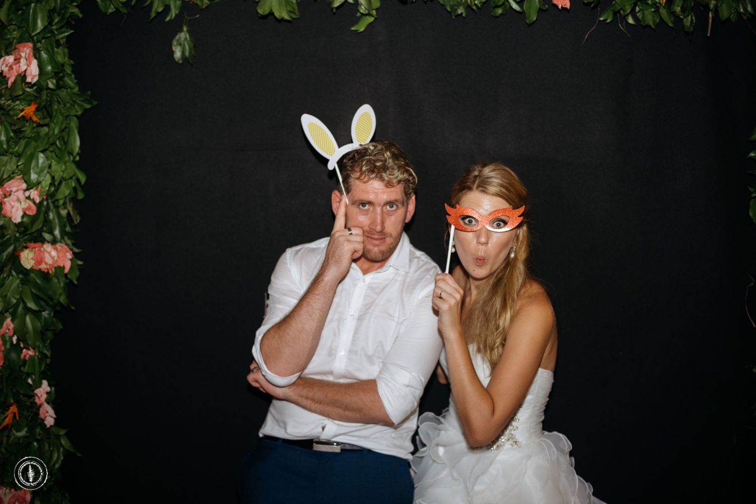 A couple wearing wedding attire and playful accessories pose with props against a dark backdrop.