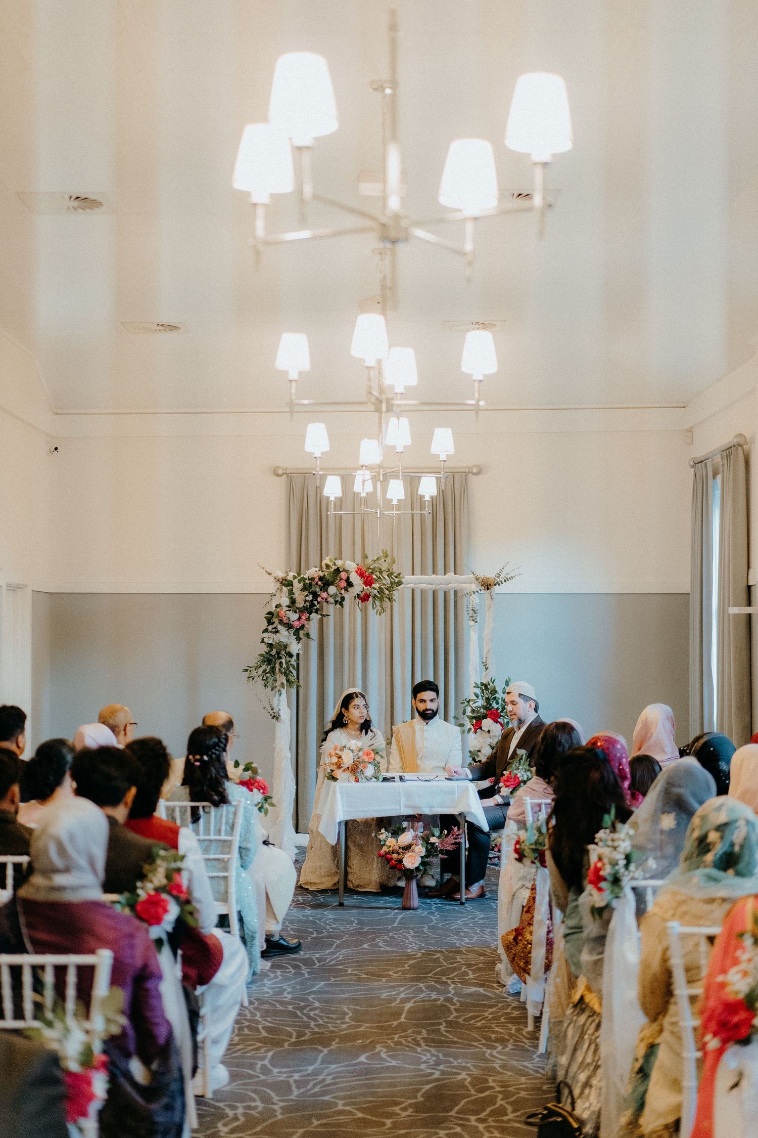 Wedding reception hall with modern chandeliers and guests seated in decorated white chairs.