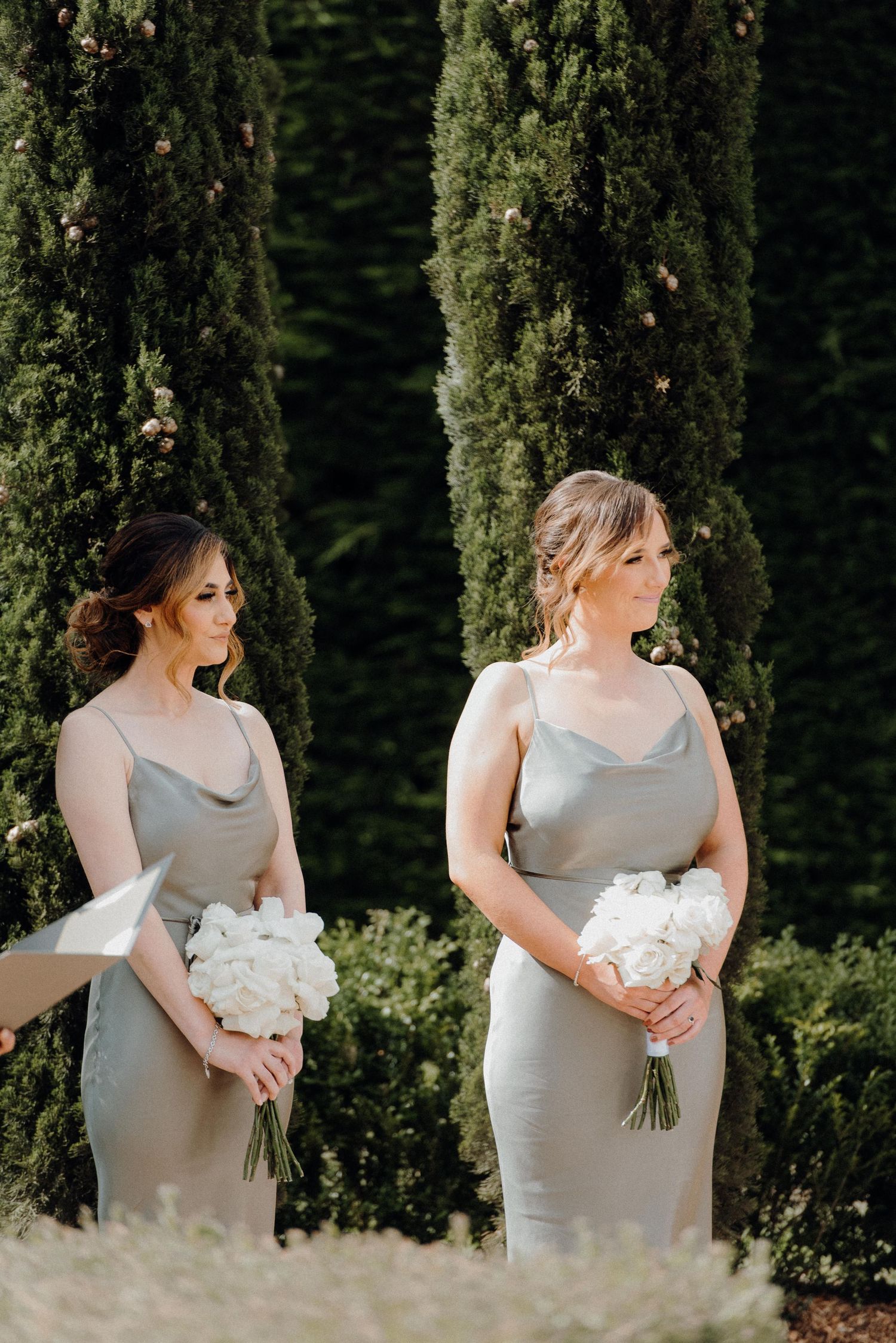 Two attendants in matching gray dresses holding white bouquets during outdoor ceremony.