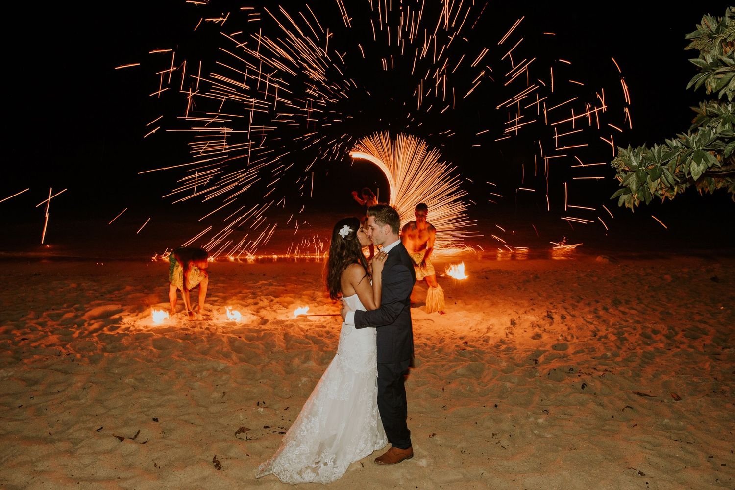 A romantic couple poses on a beach with spectacular fireworks display creating shimmering patterns behind them.