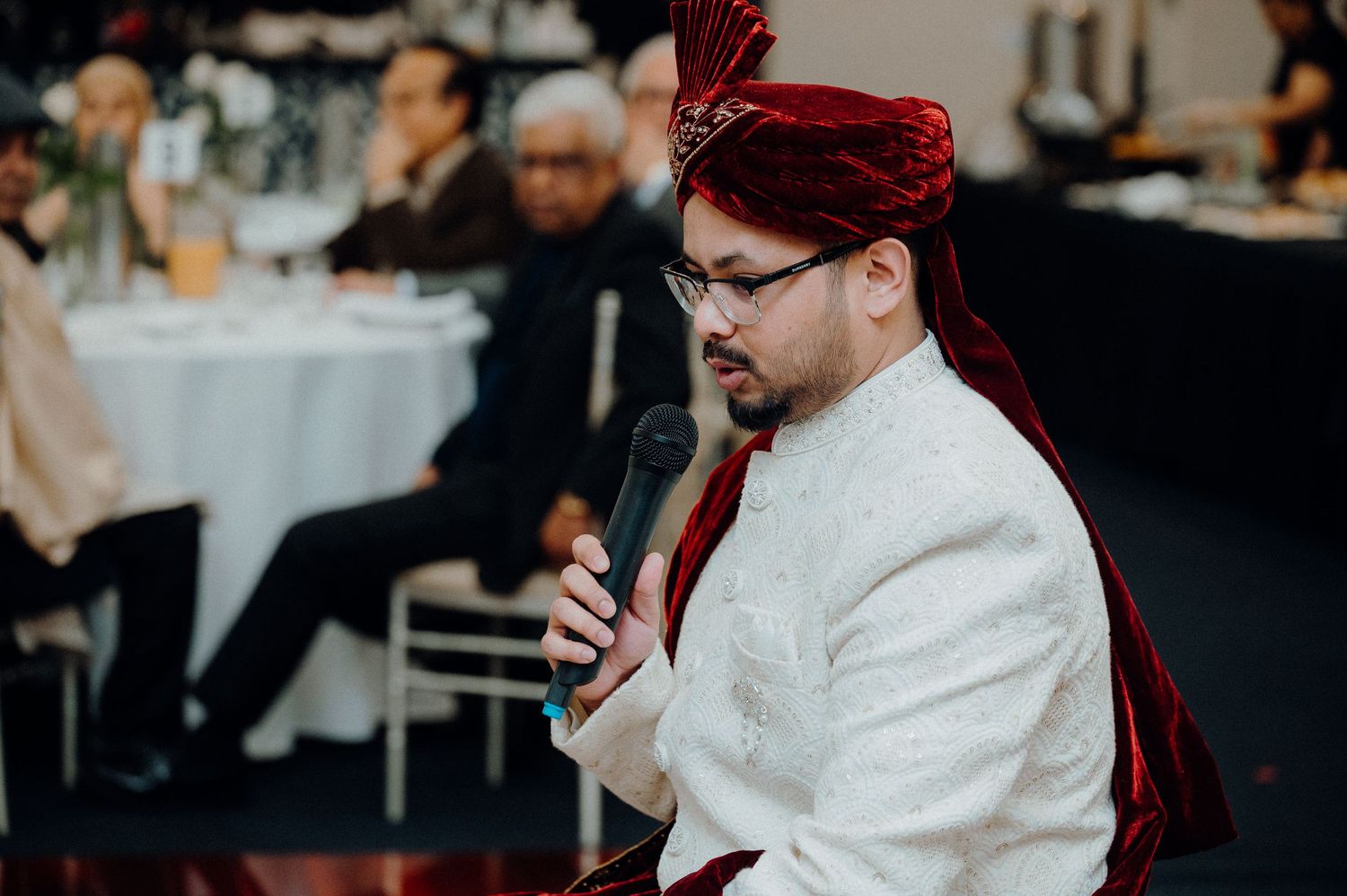 Speaker in white sherwani and red turban addressing guests at formal event.