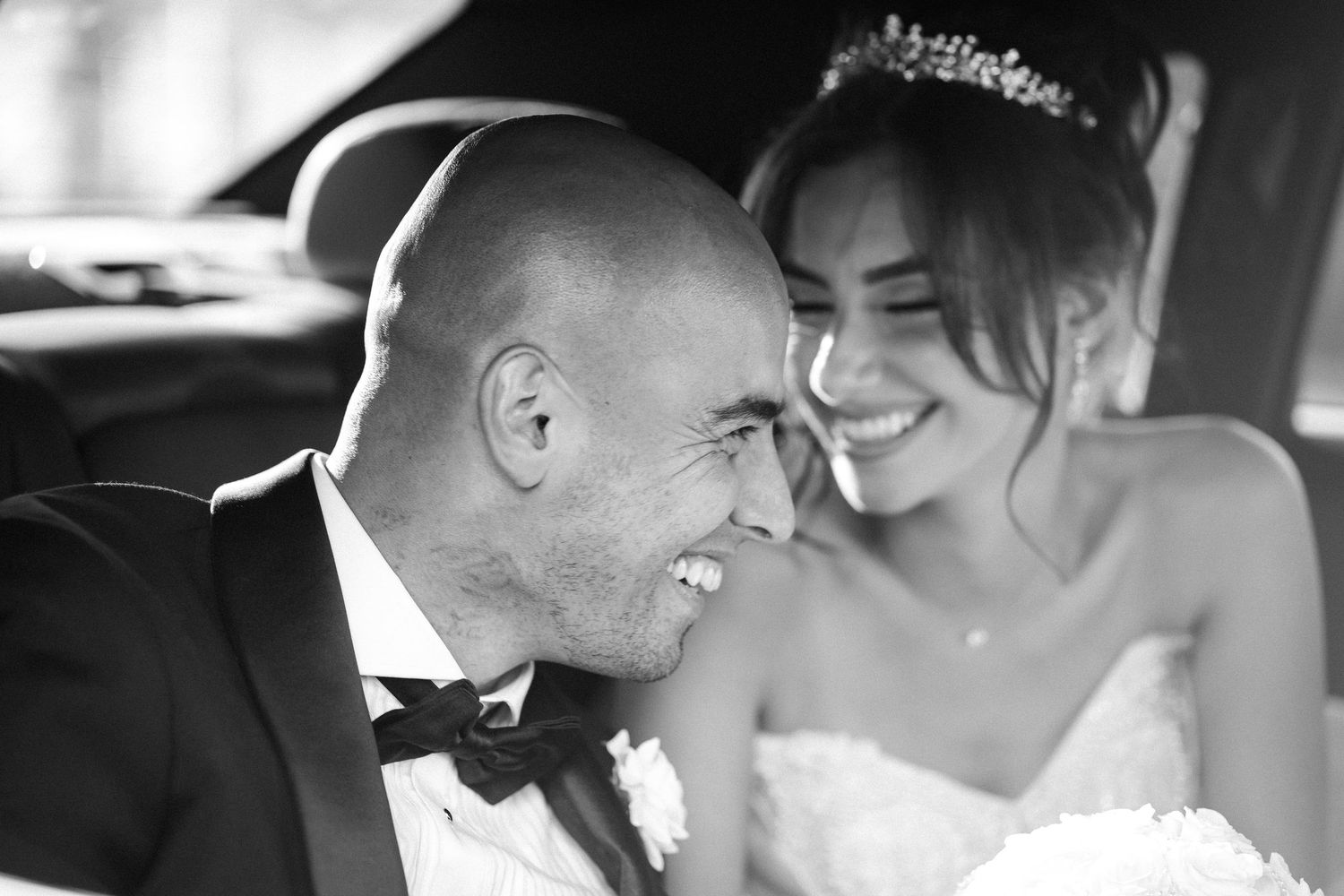 Black and white photo of smiling bride and groom sharing joyful moment in wedding car.