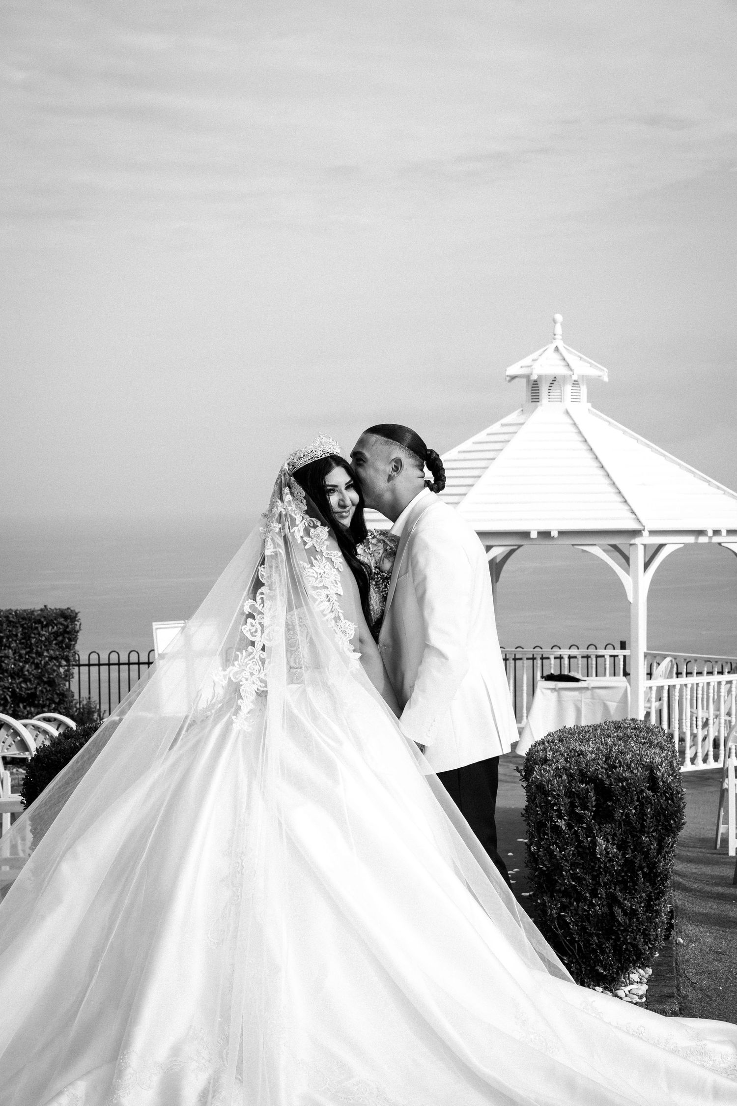 Black and white photo of a bride in a flowing gown and groom embracing near a white gazebo.