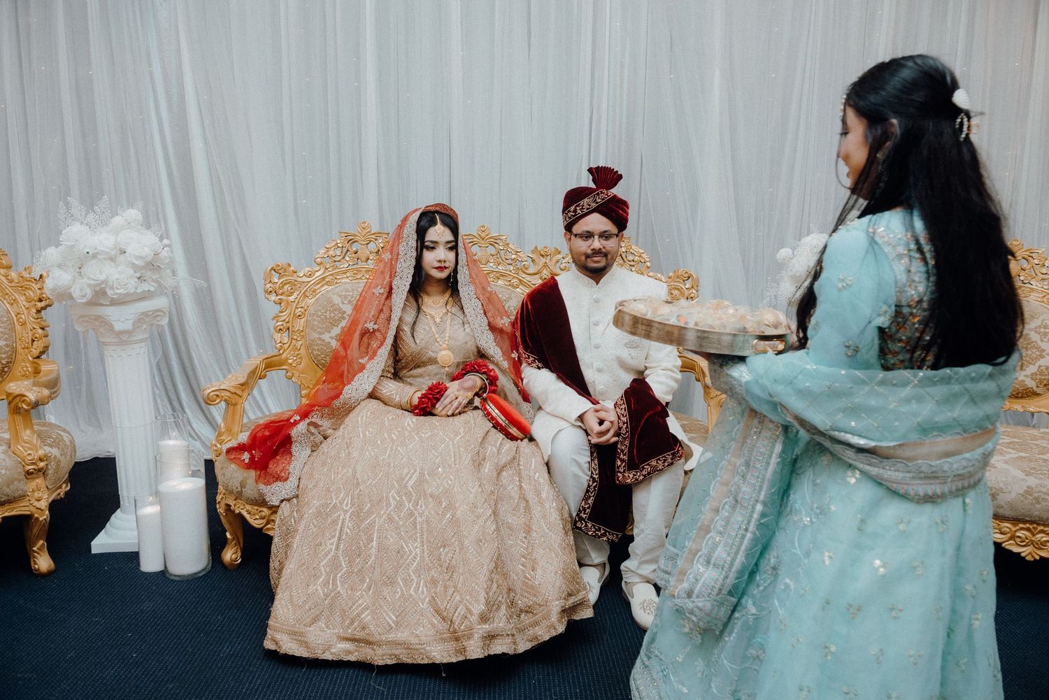 Wedding ceremony scene with participants in traditional South Asian attire seated on decorative furniture.