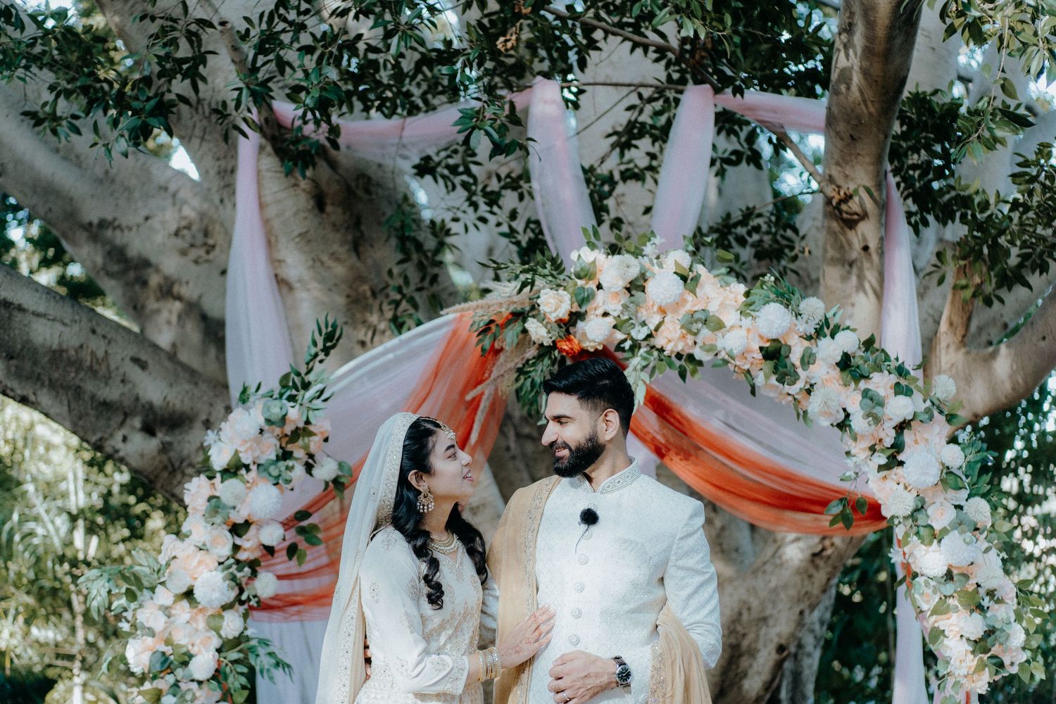 A romantic wedding ceremony under a floral arch with white and coral draping surrounded by trees.