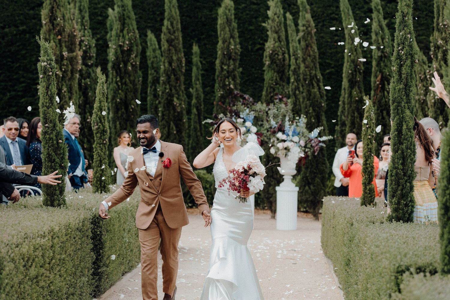 Newlyweds walking down a cypress-lined garden path while wedding guests celebrate in the background.