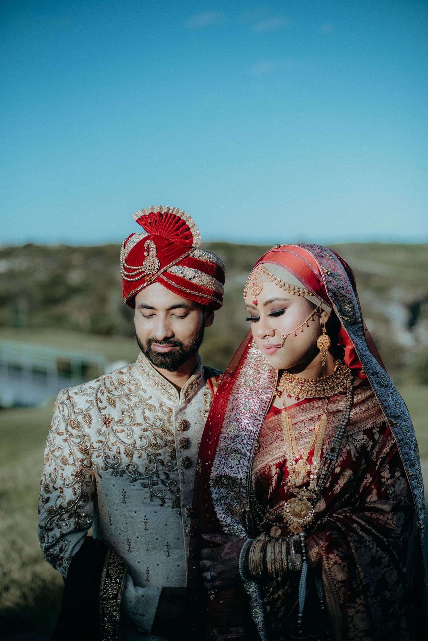 Indian wedding couple in traditional red and gold bridal attire outdoors under blue sky.