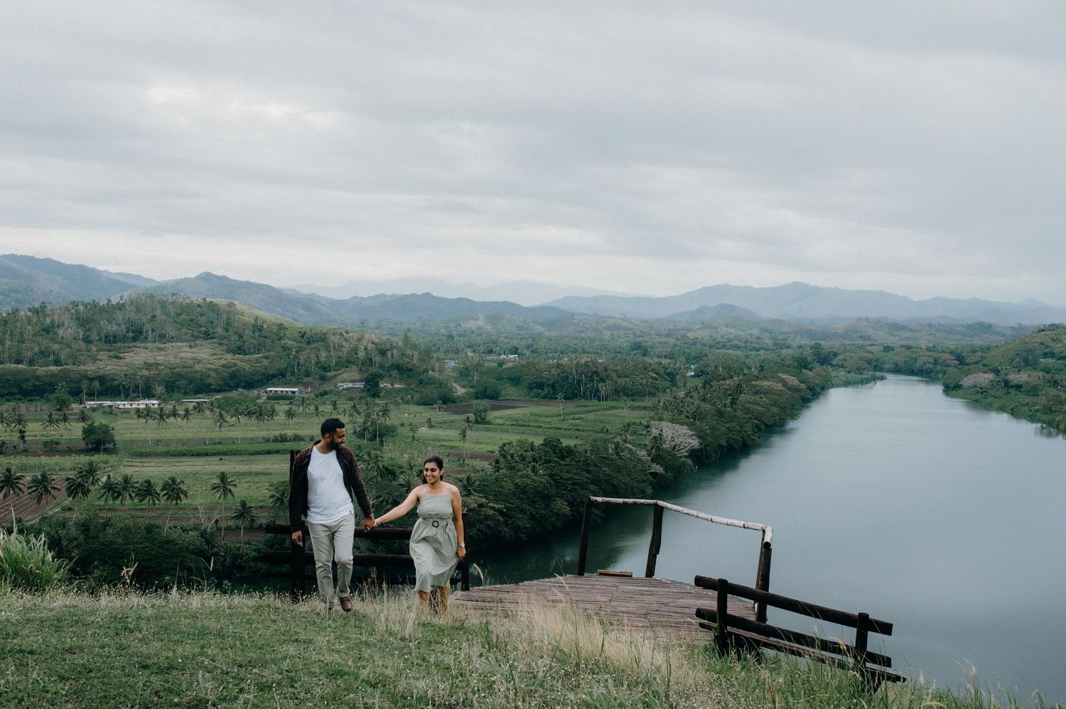 Two people hold hands while standing on a hilltop overlooking a meandering river.